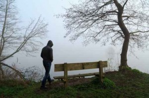 Depressed man aproaching a bench at a lake on a foggy day.